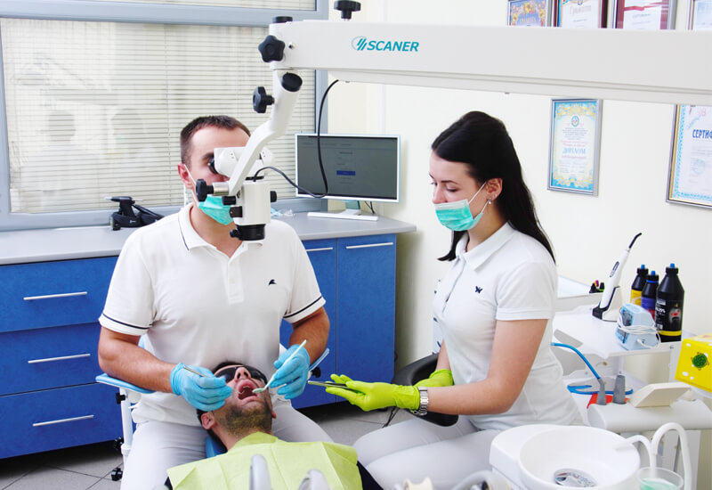 Стоматолог-терапевт кініки StomaLine працює з мікроскопом, асистент подає йому інструмент